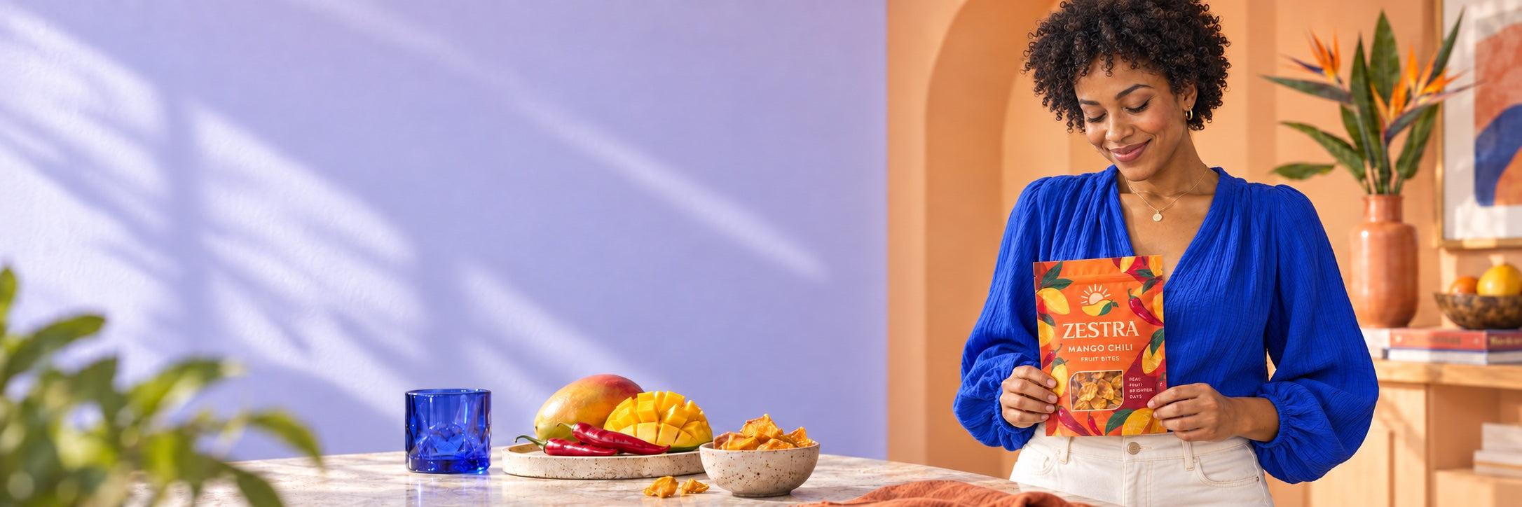 Woman in a cobalt-blue blouse looking down at a Zestra Mango Chili pouch, holding its sides naturally at waist level in a colorful studio